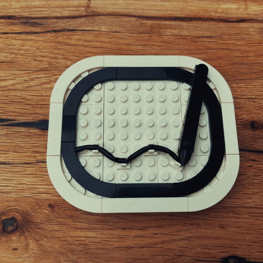 Das Logo von notiz.blog als Bricks-Modell aus schwarzen und weißen Steinen auf einem Holztisch. Eine weiße Grundplatte mit abgerundeten Ecken trägt einen schwarzen, abgerundeten Rahmen. Darin liegt ein schwarzer Stift, der unten eine geschwungene Linie zieht.