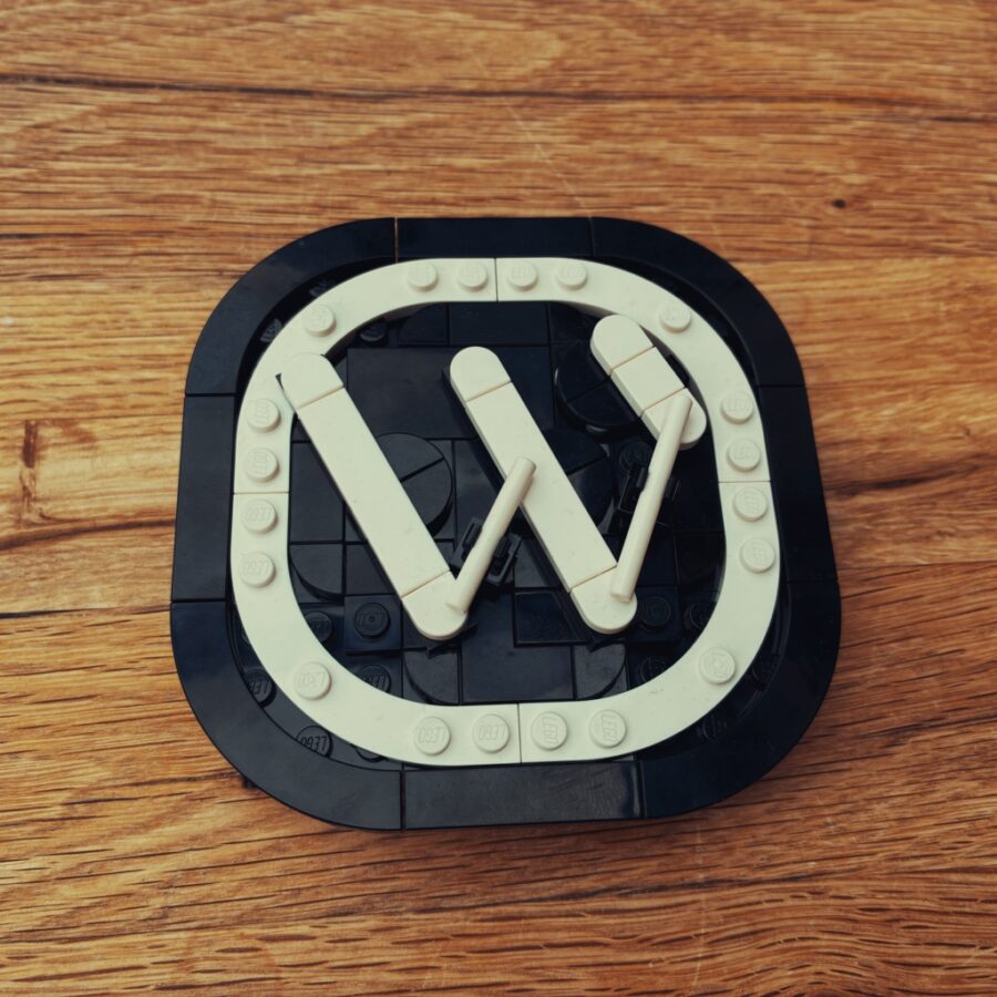 Das WordPress-Logo als Bricks-Modell aus schwarzen und weißen Steinen auf einem Holztisch. Eine schwarze Grundplatte mit abgerundeten Ecken trägt einen weißen, abgerundeten Rahmen aus Steinen mit Noppen. In der Mitte steht ein großes weißes W aus flachen, abgerundeten Teilen.